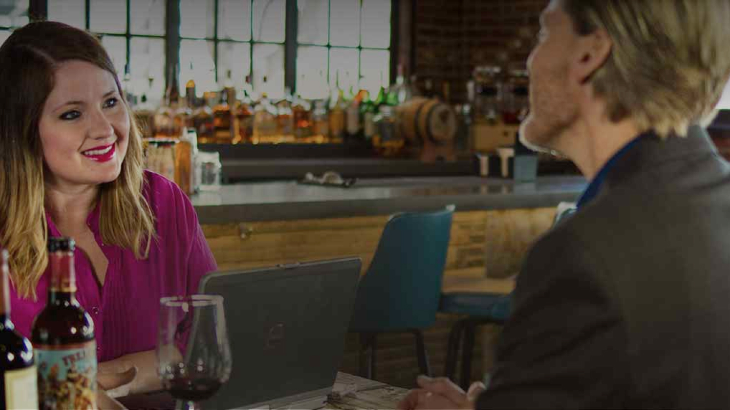 A woman in a vibrant pink blouse smiles engagingly at a man in a suit at a wine bar, with her laptop open, suggesting a friendly business meeting.