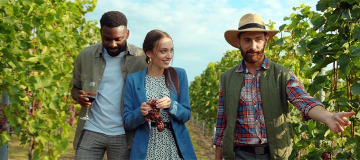Group of people in a vineyard with one holding a glass of wine and a woman holding a cluster of grapes